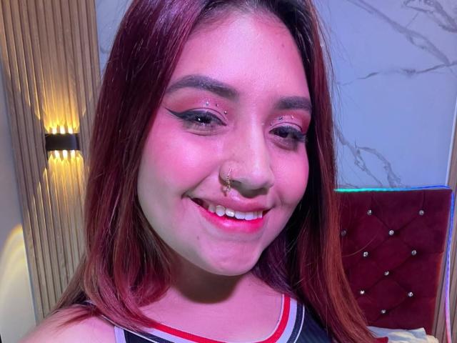 ValmontAlessia with red hair, smiling, pink eye makeup, nose ring, and septum piercing, wearing a black and red top, showing her pink lips and white teeth.