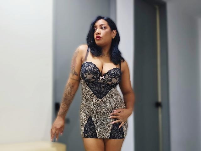 Nahirr, com cabelo preto, vestindo um vestido de renda e estampa de leopardo, exibindo seus seios grandes e o braço tatuado, está em pé com confiança, com uma mão no quadril..