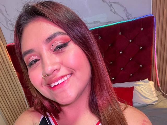 ValmontAlessia with pink eyeshadow, nose ring, and smile, showing red and black hair, pink lipstick, and white teeth. She's wearing a red and black top, looking cute.