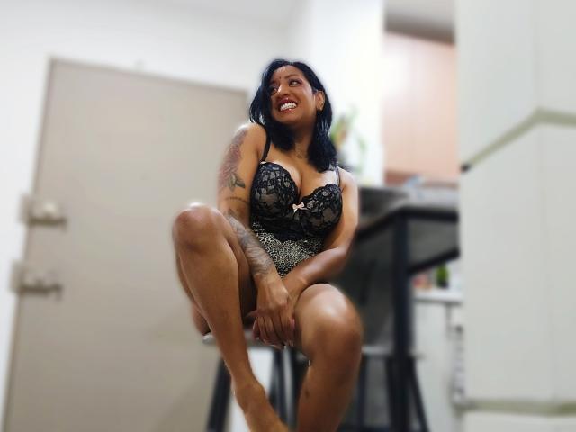 Nahirr with dark skin, black lace bra, and leopard print lingerie, squatting on a stool, smiling, showing tattoos on her arm and leg, with a cute, sexy look.