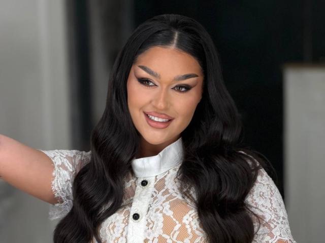 KaiaSilk, con cabello largo y ondulado de color negro, lleva una blusa de encaje blanco, está sonriendo, y lleva maquillaje con cejas oscuras y lápiz labial rosa. Tiene un brazo levantado, mostrando ligeramente sus pechos a través del encaje..