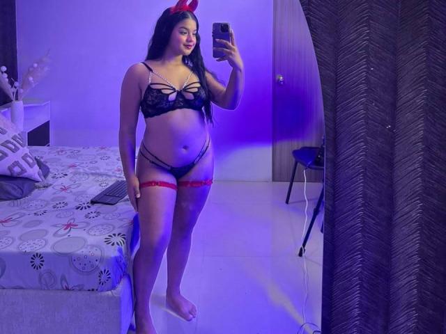 Arianna Dolche, avec de longs cheveux noirs, un nœud rouge, un soutien-gorge et une culotte en dentelle noire, une jarretière rouge, tenant un téléphone, debout devant un miroir, souriante, montrant ses seins et ses fesses..