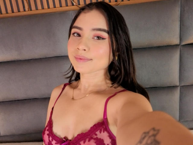 VictoriaKyros, avec ses cheveux brun foncé, son fard à paupières rose et son soutien-gorge en dentelle rouge, montrant ses petites seins et son tatouage sur le bras, souriant à l'appareil photo..