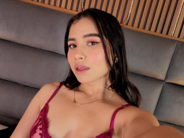 VictoriaKyros has dark hair, pink eyeshadow, and pink lipstick. She's wearing a red lace bra, showing her small tits, with a gold necklace. She's taking a selfie.