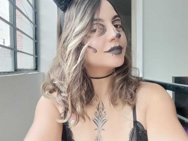 ArianaBlonde has wavy blonde hair, black cat ears, dark lipstick, and a nose ring. She's wearing a black lace top, showing a chest tattoo. Her tits are visible.