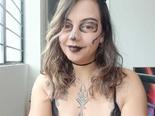 ArianaBlonde with dark lipstick, black nose ring, and makeup around her eyes, wearing a black choker and lace top, showing tattoos on her chest and arms.