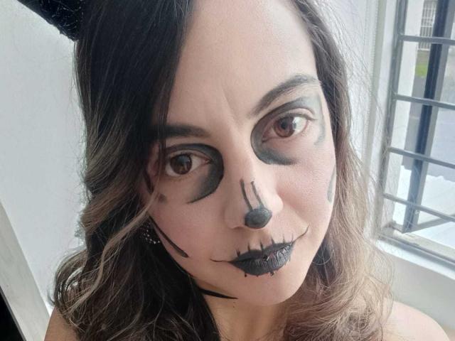 ArianaBlonde has dark, wavy hair and spooky face paint with black eyeliner, nose, and lips, looking at the camera with brown eyes. She's near a window.