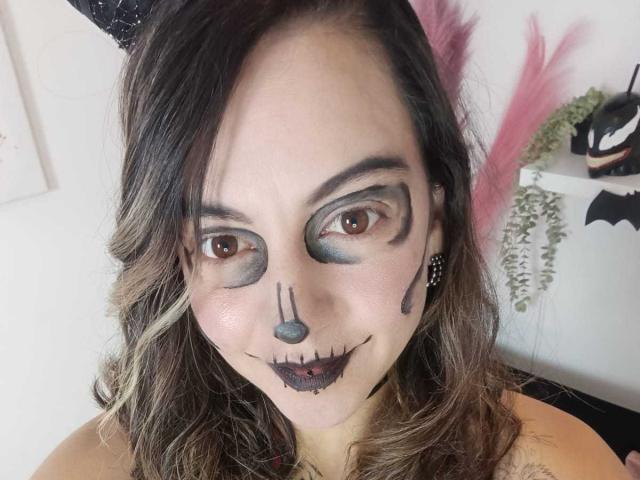 ArianaBlonde with dark makeup: gray eye shadow, black nose dot, stitched mouth, black lipstick. Brown wavy hair, black headband, pink feathers in background. She's smiling.