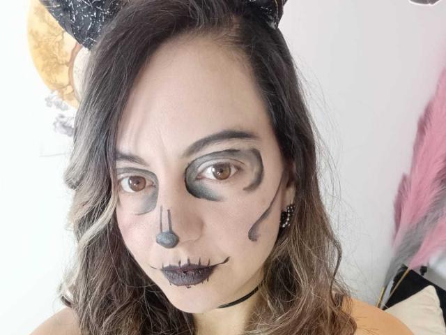 ArianaBlonde has dark makeup on her face, featuring black lines, gray eye makeup, black nose, and black lips, with long wavy brown hair and a black headband.