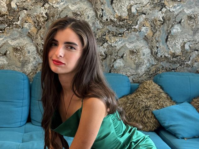 DarsiSweet with long brown hair, red lipstick, and green dress, leaning on blue couch, showing her small tits and cute ass. Fluffy pillow on right. Stone wall in background.