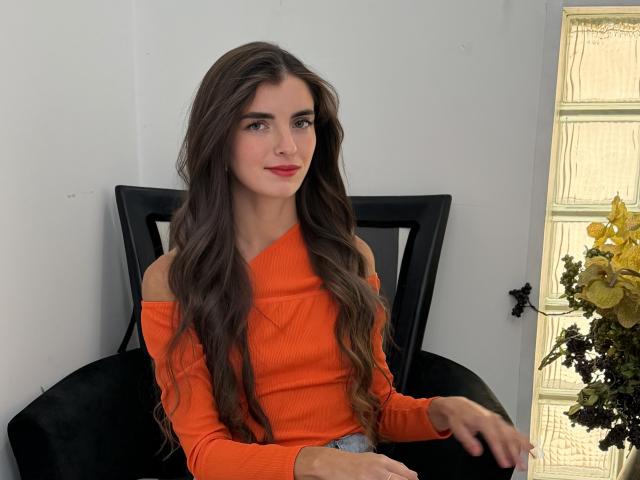 DarsiSweet with long brown hair, red lipstick, and an orange off-shoulder top, sitting on a black chair, hands resting on her lap, smiling subtly.