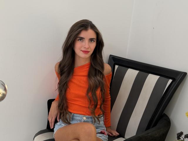 DarsiSweet with long brown hair, orange off-shoulder top, and denim shorts, sitting on a black and white striped chair, showing her ass slightly. She's smiling at the camera.