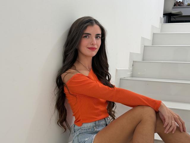 DarsiSweet, with long brown hair and red lipstick, sits on white stairs, wearing an orange off-shoulder top and denim shorts, smiling slightly. Her knees are up, hands on her thighs.