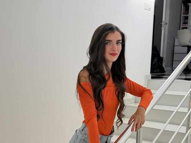 DarsiSweet with long brown hair, orange off-shoulder top, and denim jeans, leaning on a white staircase, smiling with red lipstick.