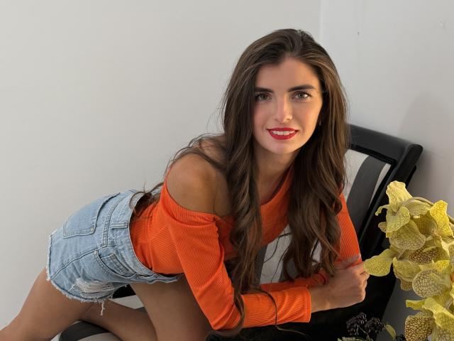 DarsiSweet with long brown hair, orange off-shoulder top, and denim shorts, leaning on a chair, holding yellow flowers, smiling with red lipstick, and showing her ass.