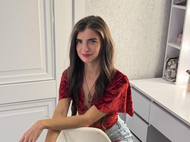 DarsiSweet with long brown hair, red velvet crop top, and denim shorts, sitting, arms crossed, wearing red lipstick, necklace, and a subtle smile, in a white room.