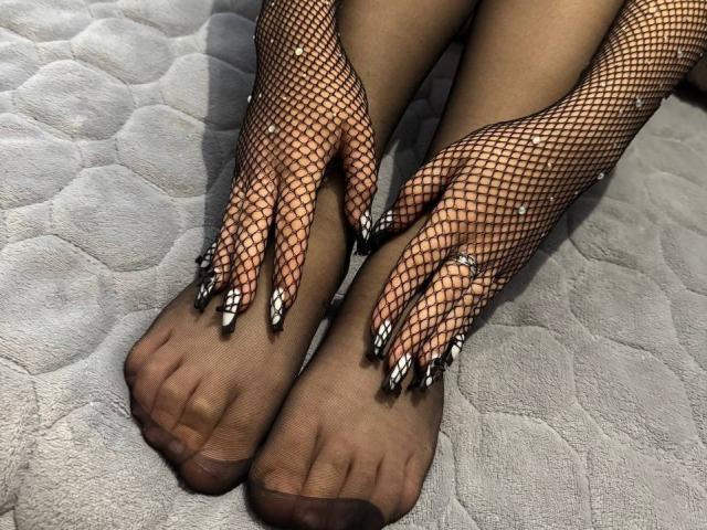  FallenAngelia's feet in black fishnet socks with long, pointed nails, white tips, and rings on her toes, lying on a gray, textured surface. Boobs are not visible.
