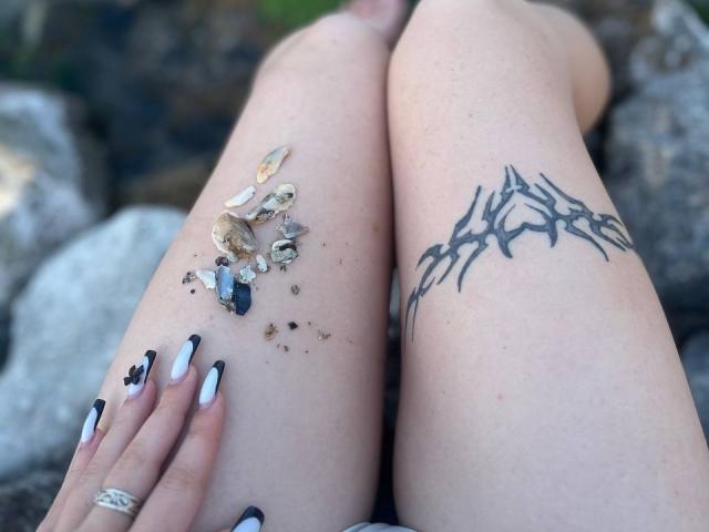 FallenAngelia's pale legs with a black tribal tattoo on the right, scattered seashells on the left, and black-white nail polish, resting on rocks. Her hand has a ring.