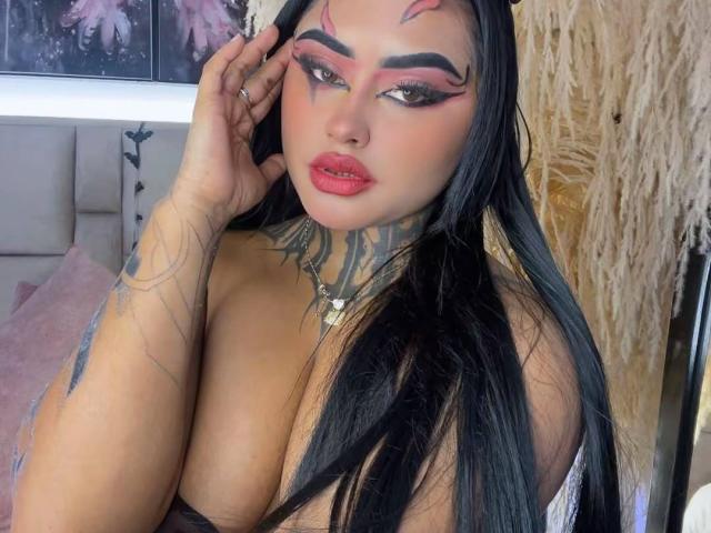 LairaHearvy with black hair, pink eyeshadow, red lipstick, tattoos, and large boobs, touching her face, wearing a necklace, in a bedroom with pink and beige decor.