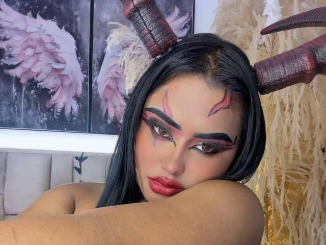 LairaHearvy with black hair, red lipstick, and dramatic eye makeup, wearing red and brown horns, resting her head on her arm, showing her bare shoulders.