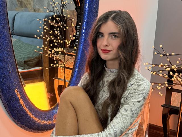 DarsiSweet with long brown hair, red lipstick, and a white patterned dress, sitting in front of a blue, glittery mirror, legs crossed, with tiny fairy lights in the background.