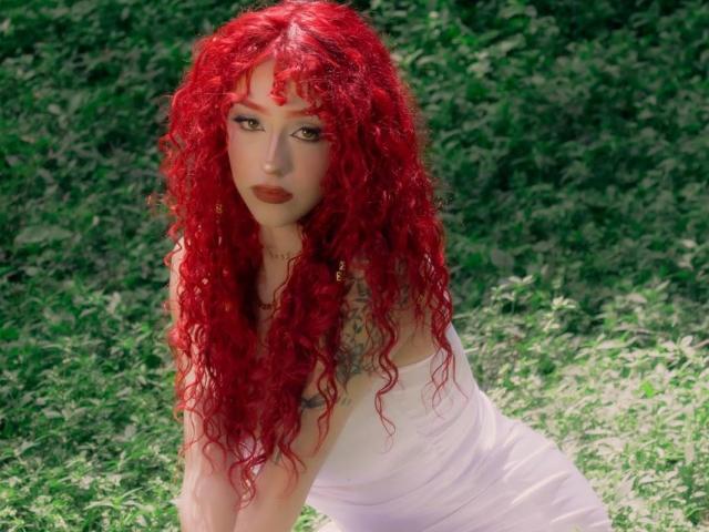 GaiaDoll con cabello rizado de color rojo brillante, vestida con un vestido blanco, inclinada hacia adelante en un área cubierta de hierba, mostrando su piel pálida y su lápiz labial rojo, con tatuajes visibles en sus brazos..