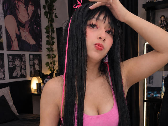 AilenRosse, avec de longs cheveux noirs, un ruban rose et un débardeur rose, faisant la moue, une main sur la tête, montrant de petites seins et du rouge à lèvres rose, dans une chambre..
