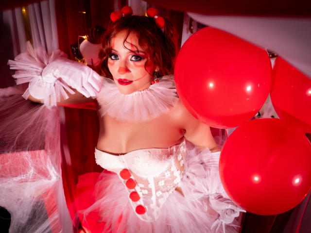 Emily Klein mit rotem Luftballon, weißer Spitzenkorsage, flauschigen weißen Handschuhen und roter, geschwollener Nase, die rote Luftballons hält und ihre Brüste zeigt, in einer rot beleuchteten, partyähnlichen Umgebung..