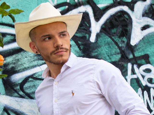 LianFerrer with a white cowboy hat, white shirt, and small shirt emblem, has a slim build, light skin, and a trimmed beard, standing in front of graffiti.