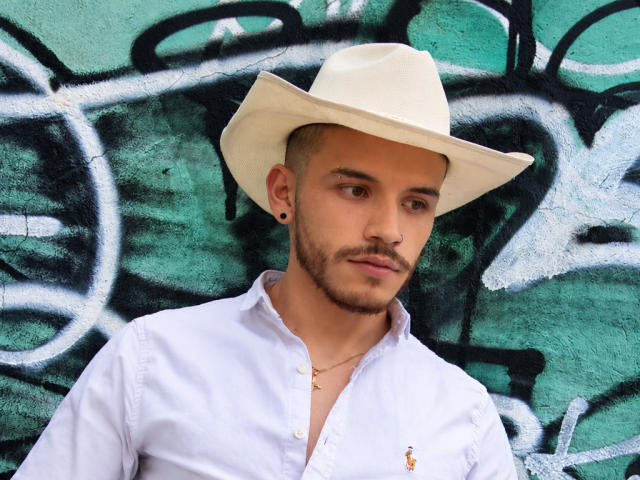 LianFerrer with a white cowboy hat, white button-up shirt, and a small gold cross necklace, sporting a trimmed beard and black ear studs, looking slightly to the side.