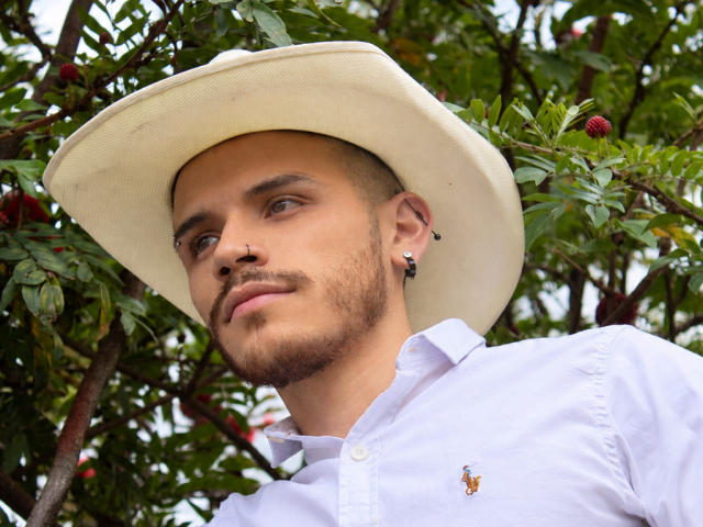 LianFerrer with a white hat, white shirt, small beard, ear piercings, and a slight smirk, looking up, surrounded by green leaves and red berries.
