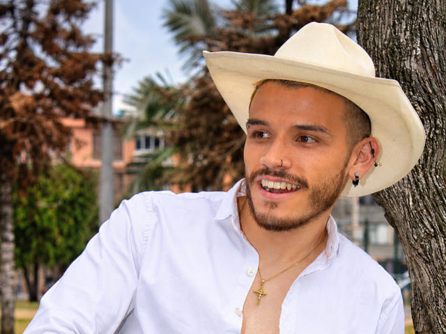 LianFerrer with a tan skin tone, wearing a white cowboy hat, white shirt, cross necklace, and black earrings, smiling with a beard and nose ring, standing by a tree.