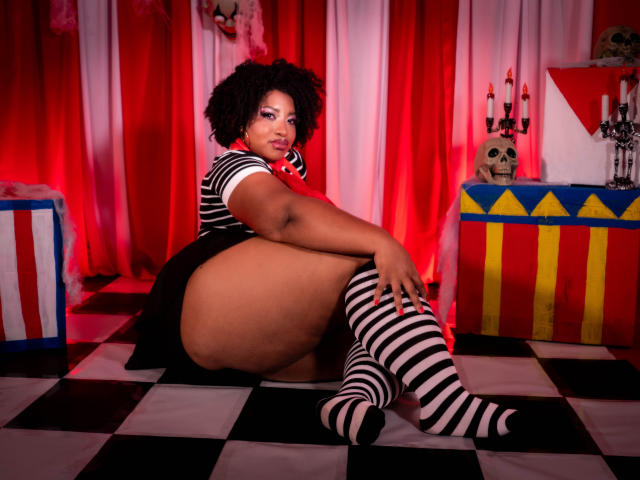 ViaraMoon with curly black hair, striped dress, and matching knee socks, sitting on checkered floor, showing big ass, looking at camera, red and white background, candles on table.