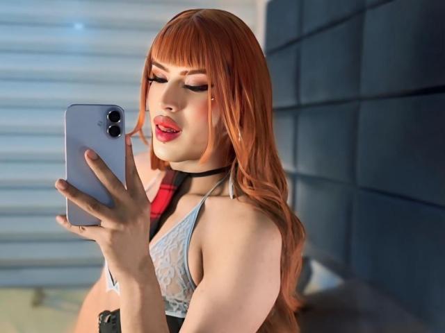 MistressNnyx, avec ses longs cheveux roux, portant un soutien-gorge en dentelle, un collier noir et du rouge à lèvres, prend un selfie avec un iPhone argenté, les yeux fermés et les lèvres légèrement entrouvertes..