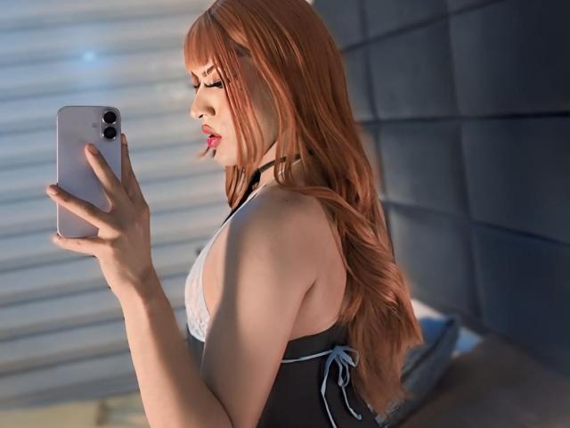 MistressNnyx, com cabelo ruivo, segurando um telefone, vestindo uma blusa preta e branca com cadarços, olhando para seu reflexo, mostrando seus pequenos seios e pele clara..