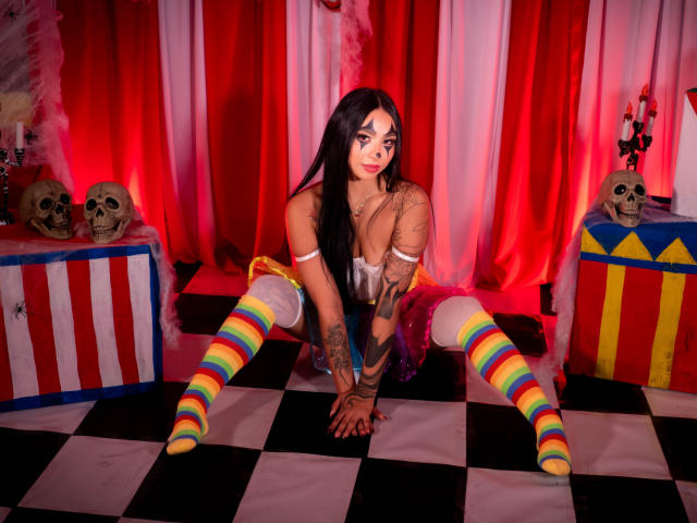 ChloeMoretti, avec ses longs cheveux noirs, sa poitrine généreuse, ses tatouages et ses chaussettes multicolores, est agenouillée sur un sol à carreaux noirs et blancs, vêtue d'un haut transparent et de brassards blancs, dans un décor éclairé en rouge..