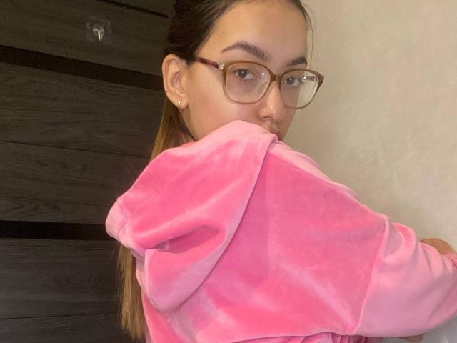RaneeDerienzo mit Brille, braunen Haaren im Pferdeschwanz, trägt eine rosa, flauschige Kapuzenjacke, beißt am Ärmel, blickt über ihre Schulter, vor einer dunklen Holzwand..