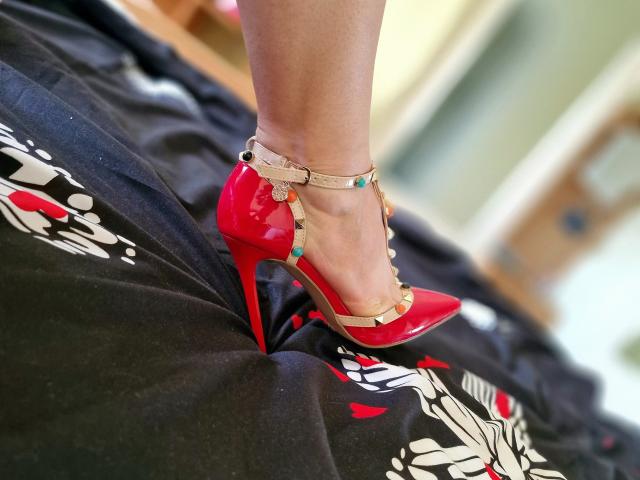 Zaccada's shiny red stiletto heels with gold straps and turquoise beads, standing on a black bed with white and red floral patterns. Her legs are slightly bent.