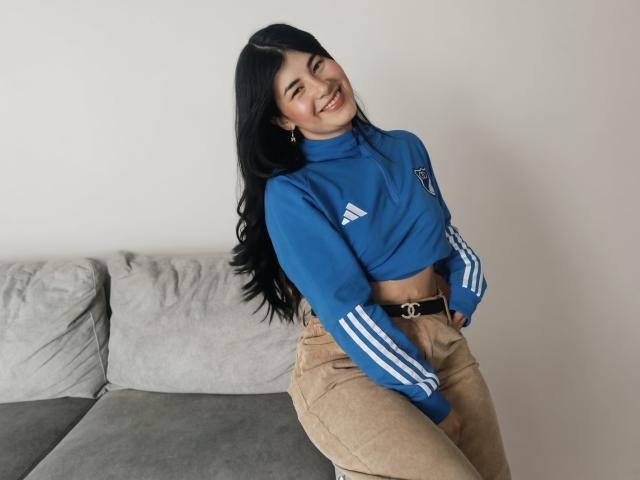 NinaHarpper with long black hair, smiling, wearing a blue Adidas crop top, tan high-waisted pants, and a black belt, sitting on a gray couch.