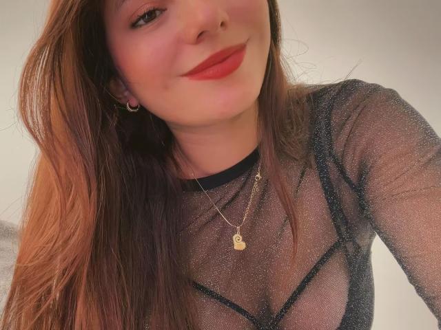 LinaFoxyHot, com cabelos castanhos longos, vestindo uma blusa preta transparente, batom vermelho e um colar dourado, sorrindo para a câmera. Seus seios são visíveis através do tecido transparente..