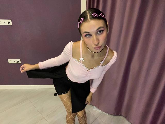 BrendaBovia with pink headband, pink top, black skirt, fishnet stockings, bending forward, pouting, showing ass, small tits, pink eyeliner, white floor, purple wall, black choker.