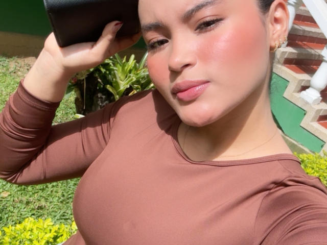 MiiaPalmer with pink blush on her cheeks, holding a black phone, wearing a brown long-sleeve top, showing her large boobs, smiling with pink lipstick, in a sunny outdoor setting.