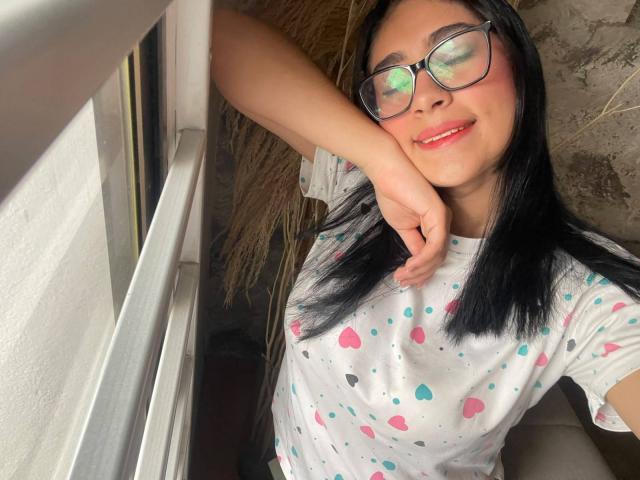 NinaRouse with black hair, glasses, pink lipstick, smiling, hand resting on window, wearing white shirt with pink and blue dots, looking out. Her tits are small.