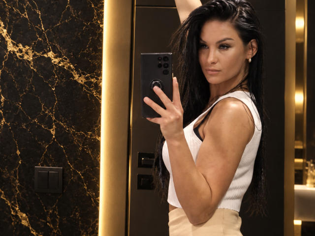 IvyMidnight with long black hair, wearing a white tank top, holding a phone, showing muscular arms, and a confident expression, standing in a modern, dark-walled bathroom.
