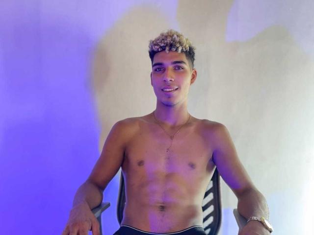 Shirtless SantiaGodiazz with curly blonde hair, tan skin, and a slim, toned body. He's smiling, wearing a necklace and wristwatch, with blue and white lighting.
