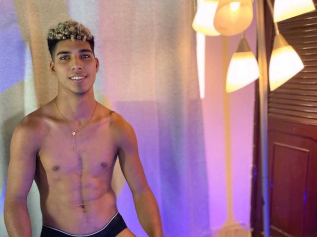 SantiaGodiazz, shirtless with curly blonde hair, medium brown skin, small chest, and visible abs, wearing black briefs, smiling, standing next to a lit floor lamp.