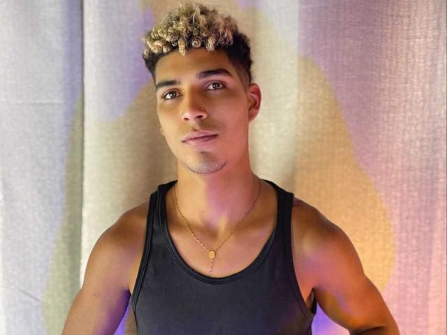 SantiaGodiazz has curly blonde and brown hair, tan skin, brown eyes, a black tank top, and a gold necklace with a small circular pendant. He's looking slightly to the side.