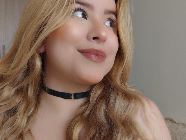 AshleySofia, con cabello rubio y ondulado, piel clara y una sonrisa sutil, lleva un collar negro, mira hacia arriba con labios brillantes y maquillaje suave..