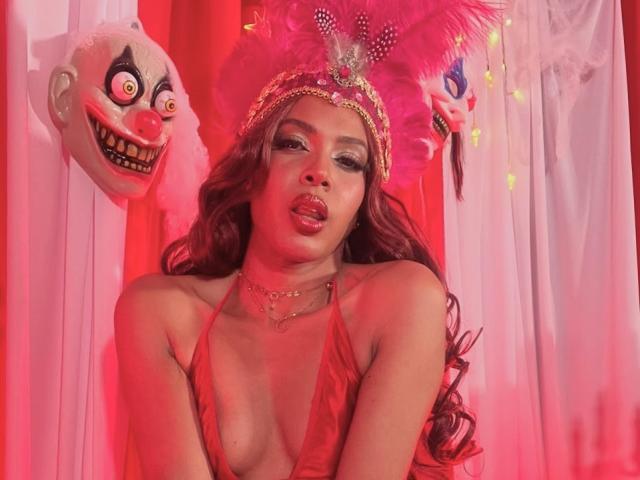 CamilaWest con rossetto rosso, capelli castani ondulati e un copricapo rosa con piume, indossa un abito rosso a scollo basso che mette in mostra il suo seno piccolo, e collane dorate, con maschere da clown sullo sfondo..
