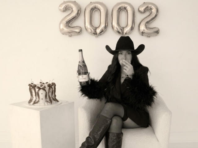 JennyLawry with long dark hair, black cowboy hat, fur coat, black dress, and knee-high boots, holding a champagne bottle, sitting in a chair, sucking on her finger.